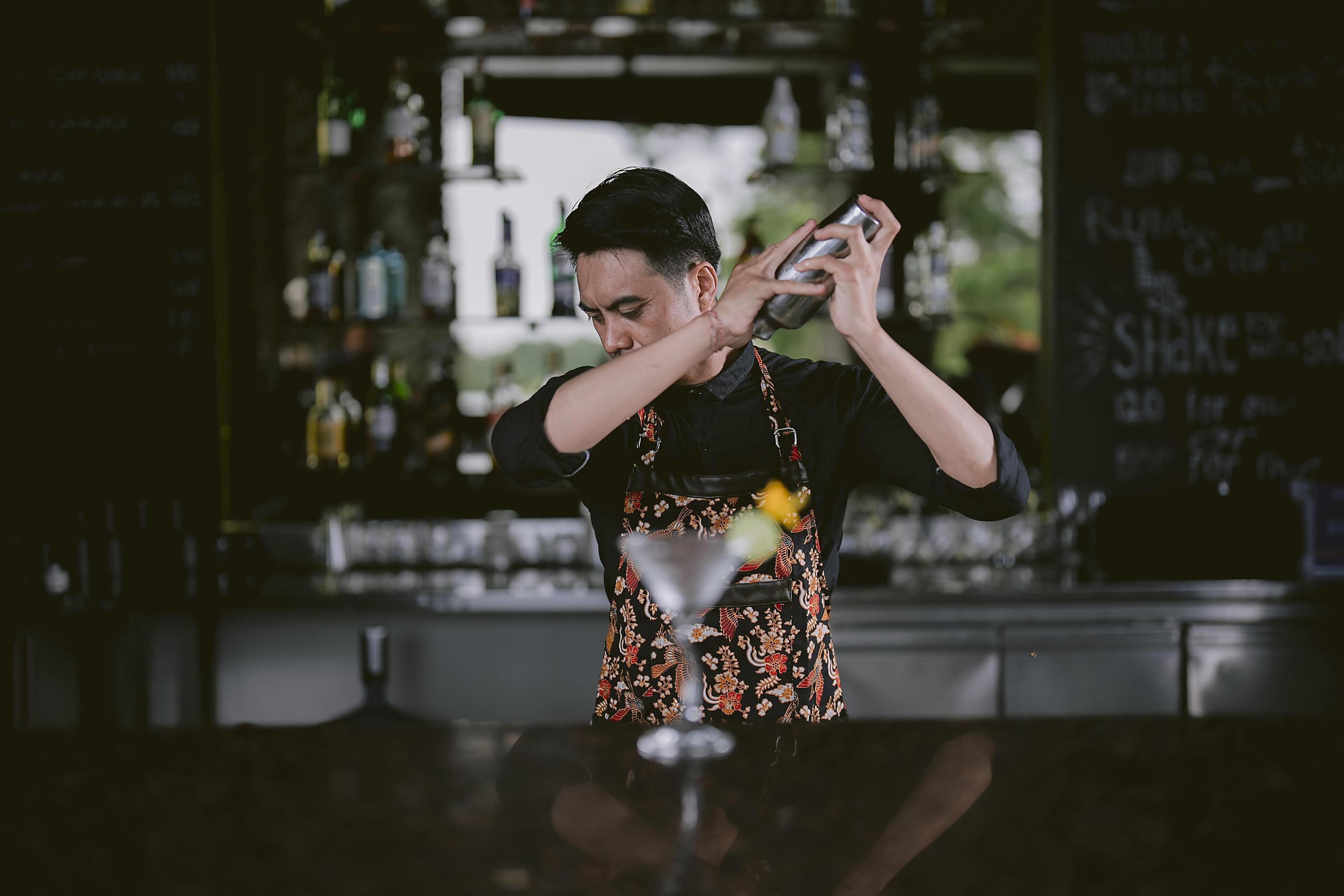 A bartender preparing a drink at a bar with shelves of bottles in the background.