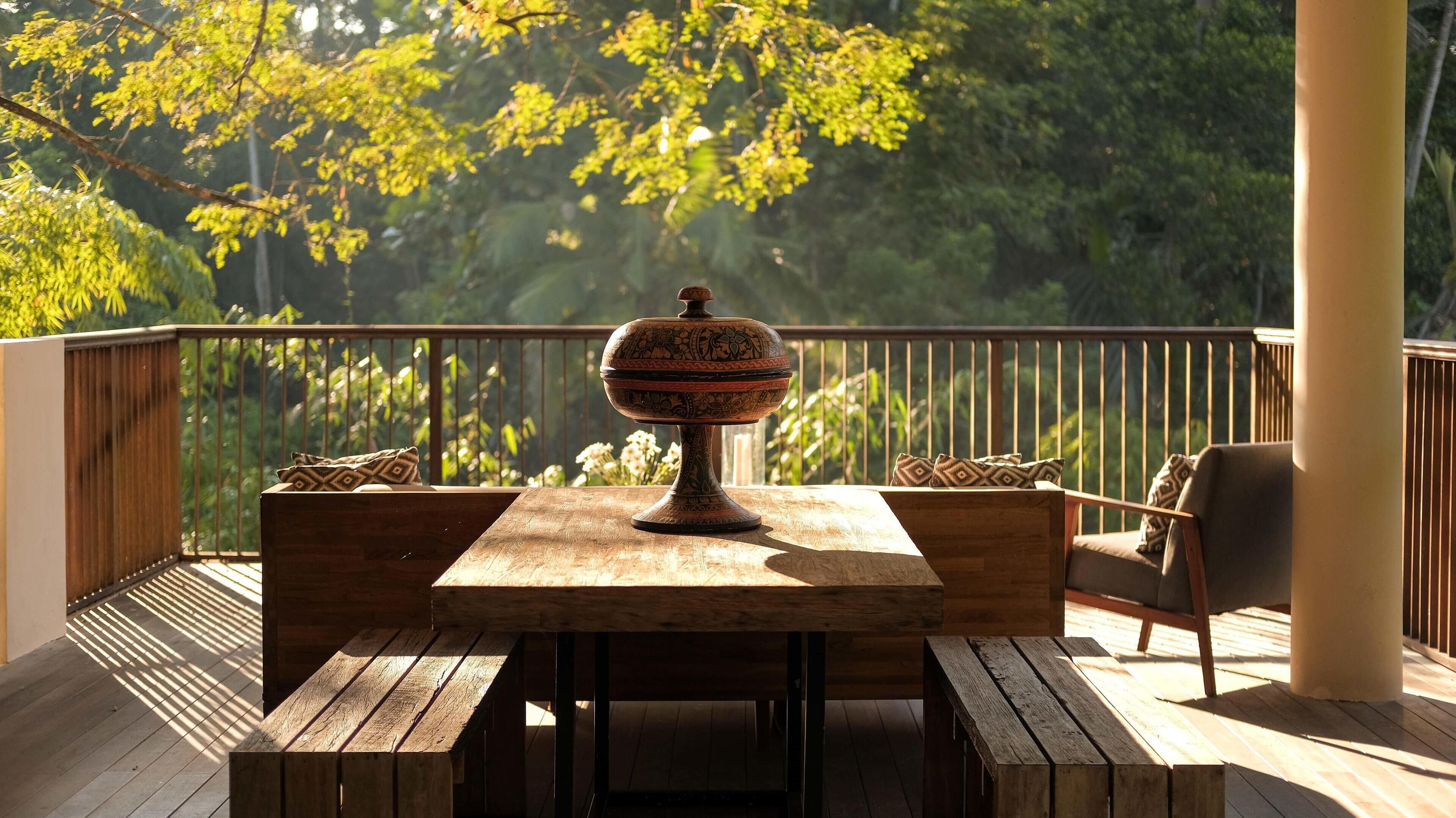 Una terraza de madera con una mesa, sillas, un cuenco decorativo y una barandilla con vistas a un área boscosa.