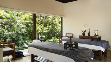 A room with two beds, a wooden bench, a small table with a tray, and a large window overlooking a lush garden.