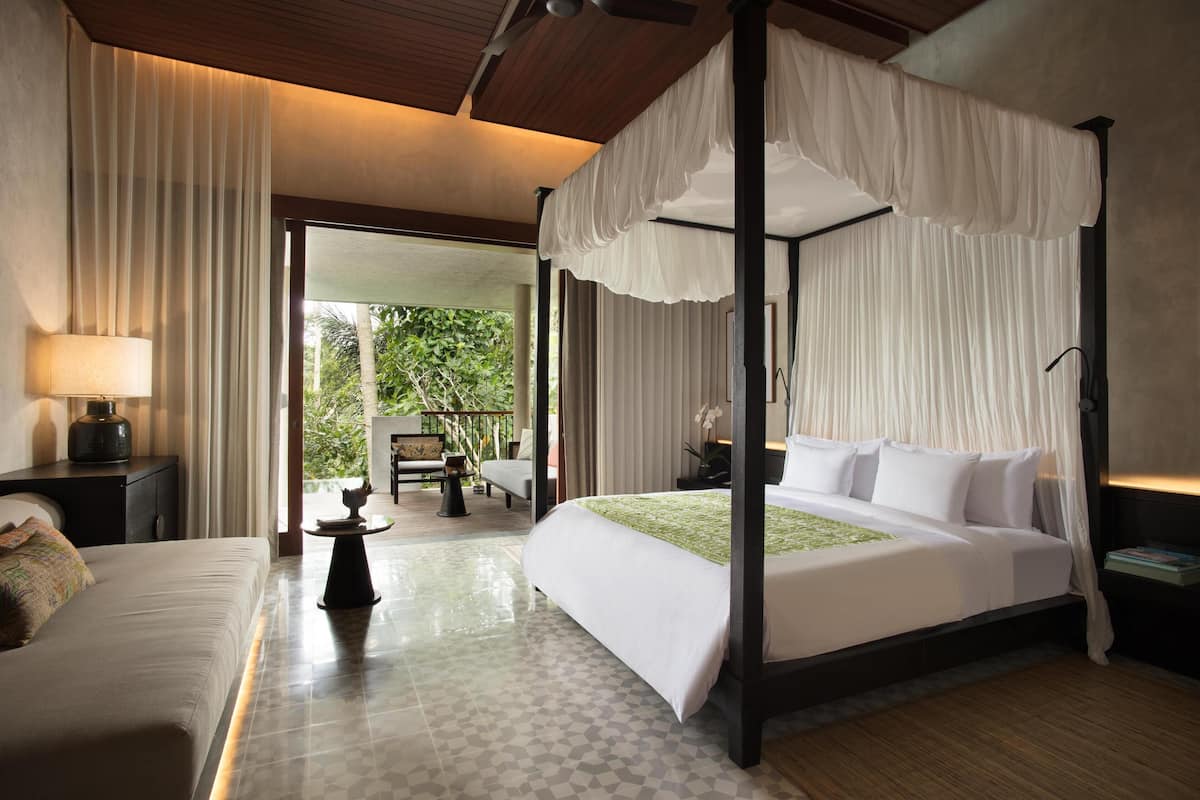A four-poster bed with white bedding, a bedside table, a lamp, and a view of a balcony with outdoor furniture.