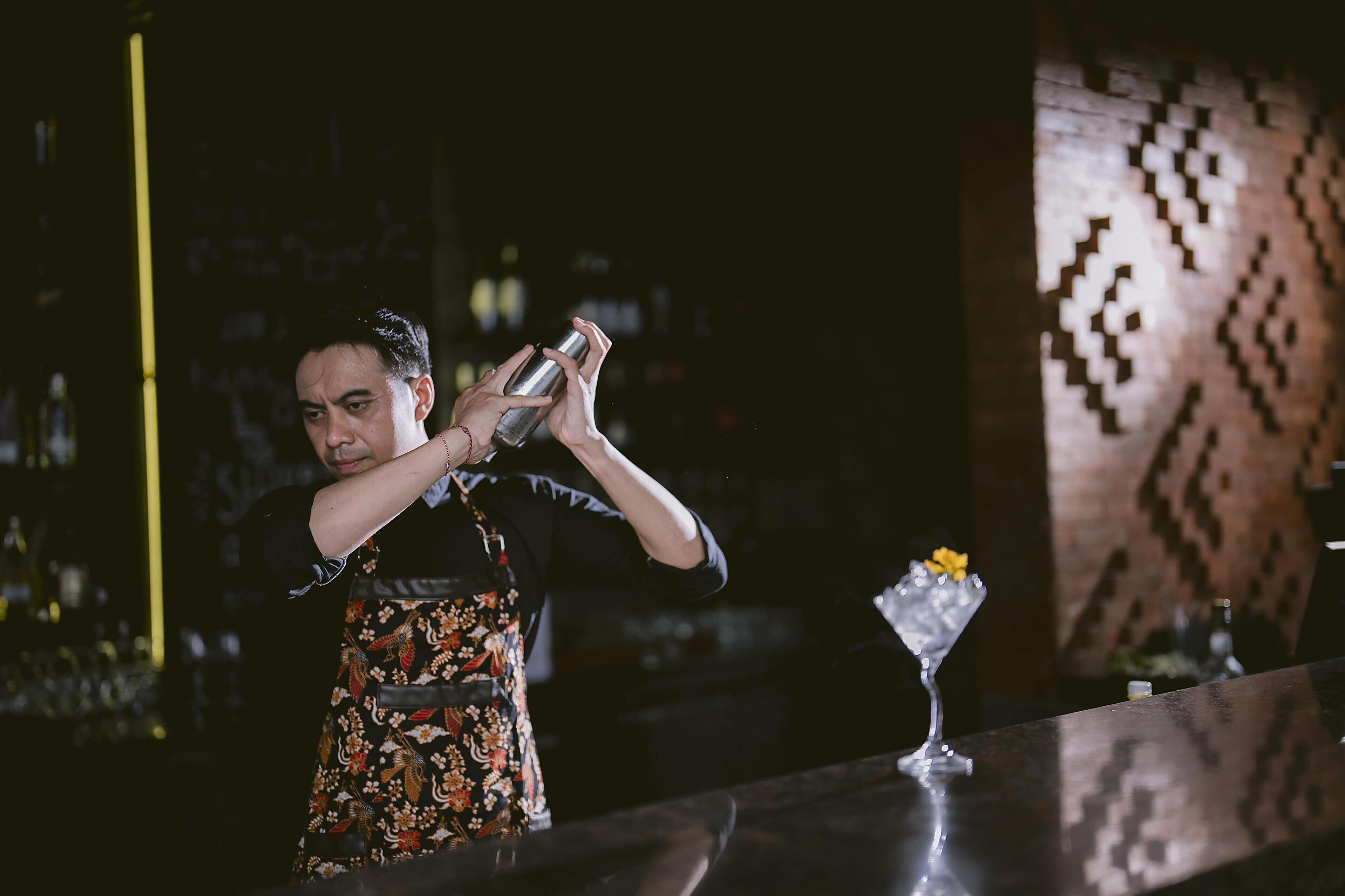 A bartender pouring a drink at a bar with a patterned wall and a decorative glass on the counter.
