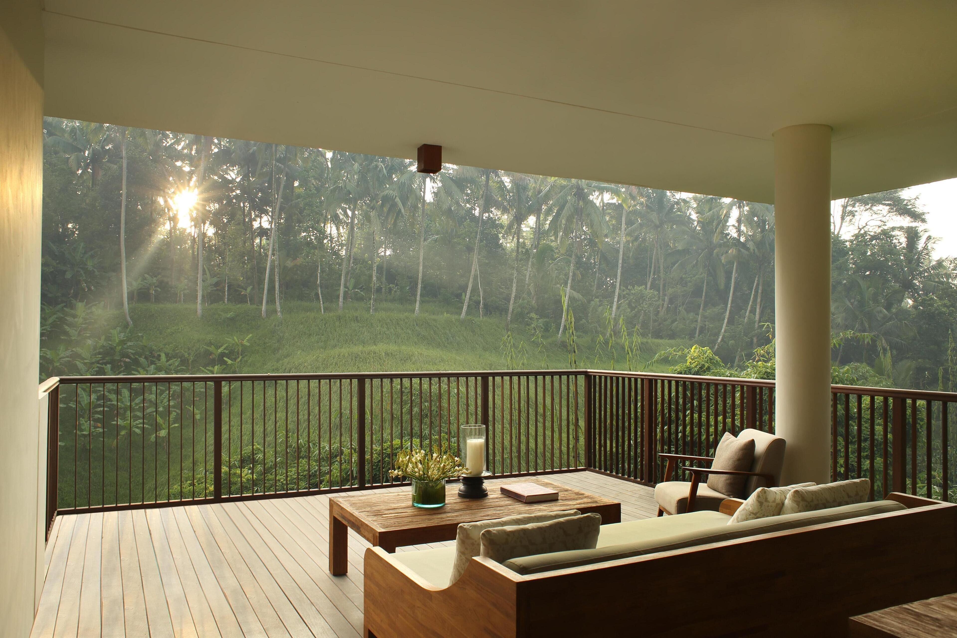 A wooden deck with a sofa and a table, overlooking a lush green landscape with tall trees and a clear view of the sun shining through.