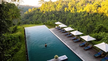 Uma piscina infinita retangular com uma pessoa nadando, cercada por espreguiçadeiras e guarda-sóis, ao fundo uma floresta exuberante.