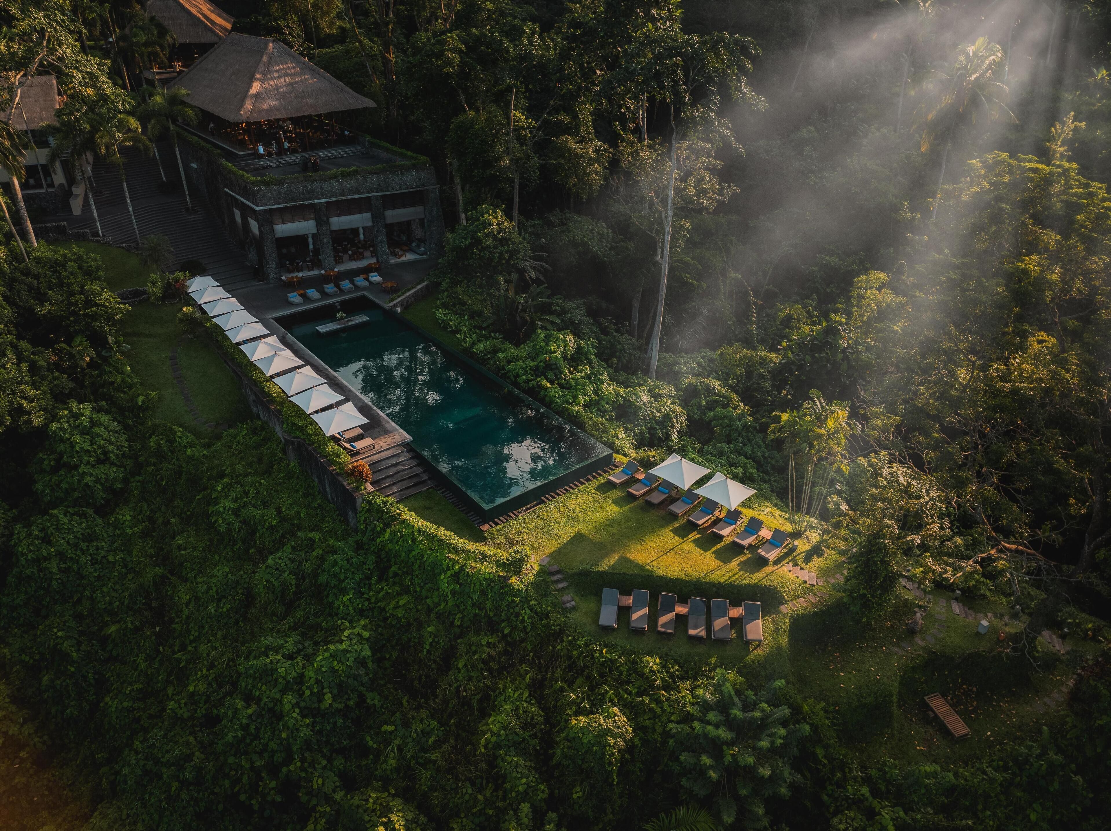 Eine Vogelperspektive zeigt ein Resort mit Schwimmbad, Liegestühlen und einem Gebäude mit einem Reetdach.