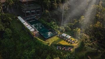 Eine Vogelperspektive zeigt ein Resort mit Schwimmbad, Liegestühlen und einem Gebäude mit einem Reetdach.