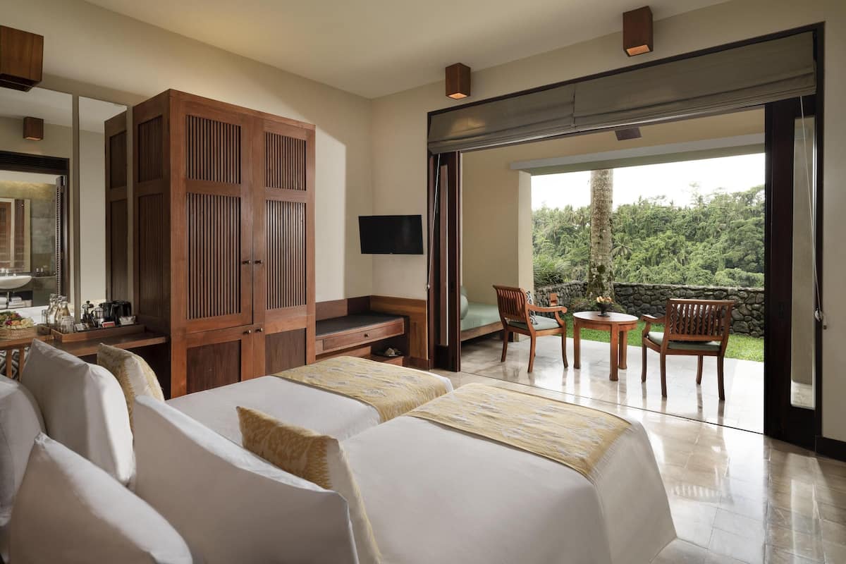 A hotel room with two beds, a wooden wardrobe, a television, a balcony with chairs, and a view of greenery.