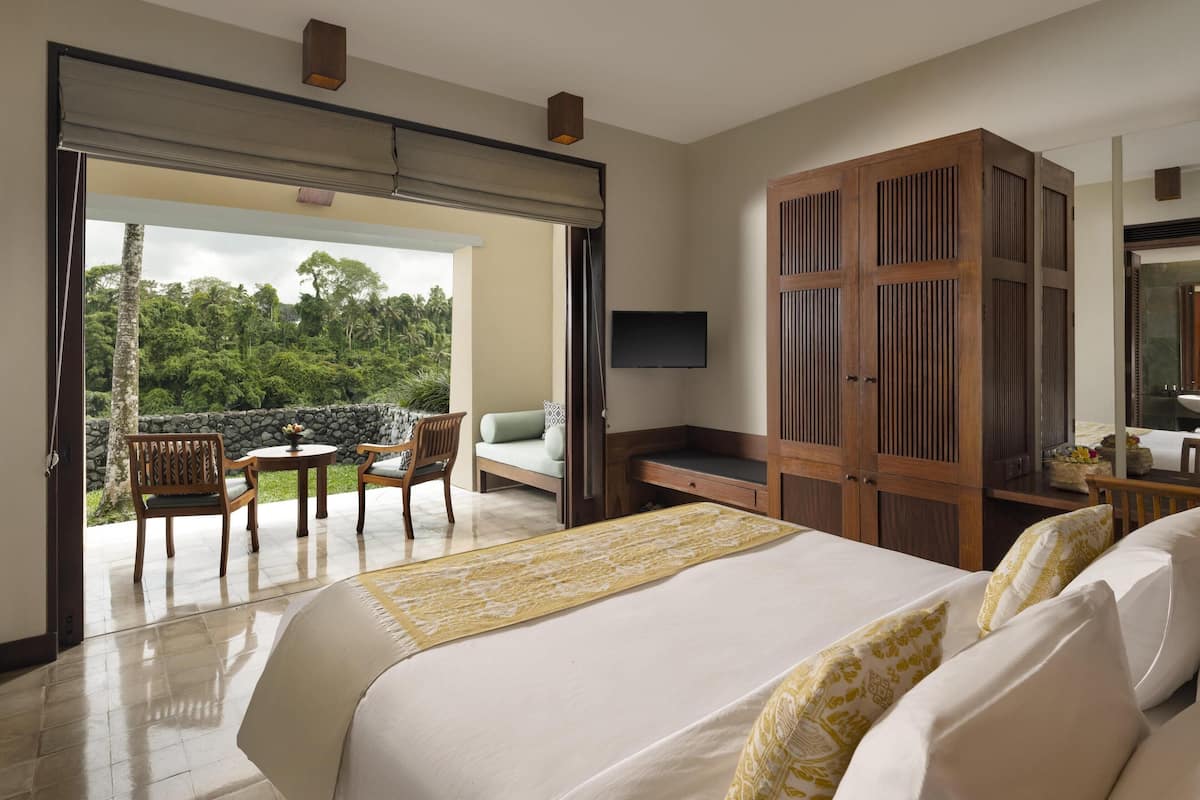 A hotel room with a bed, a desk, a chair, and a television. There is a balcony with a view of the forest.