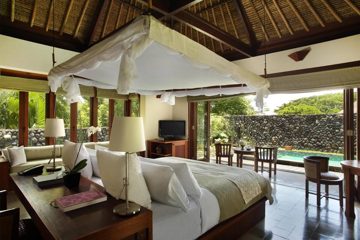 A bedroom with a canopy bed, a TV, a desk with a lamp, and a view of a swimming pool and greenery.