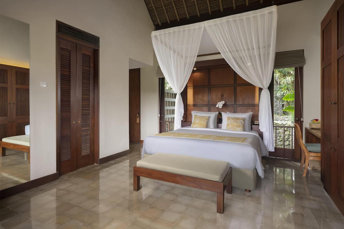 A bedroom with a large bed, a bench, wooden doors, and a view of greenery outside.