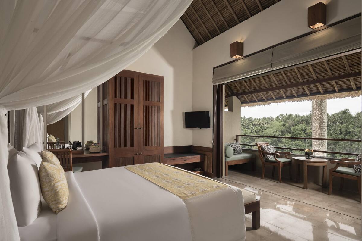 A bedroom with a large bed, a wooden wardrobe, a TV mounted on the wall, and a balcony with seating.