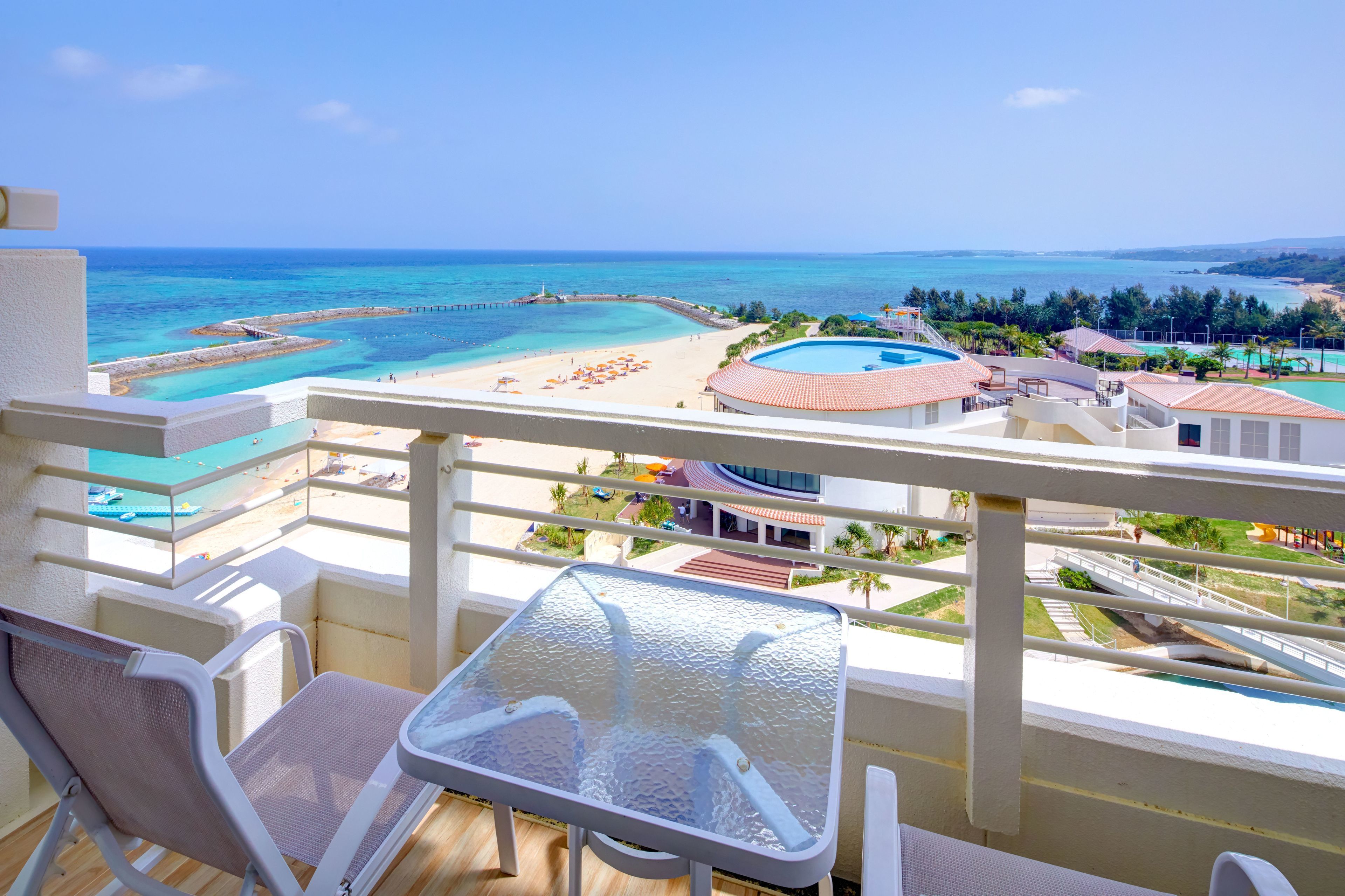 A balcony with a view of a beach, a pool, and a clear sky.