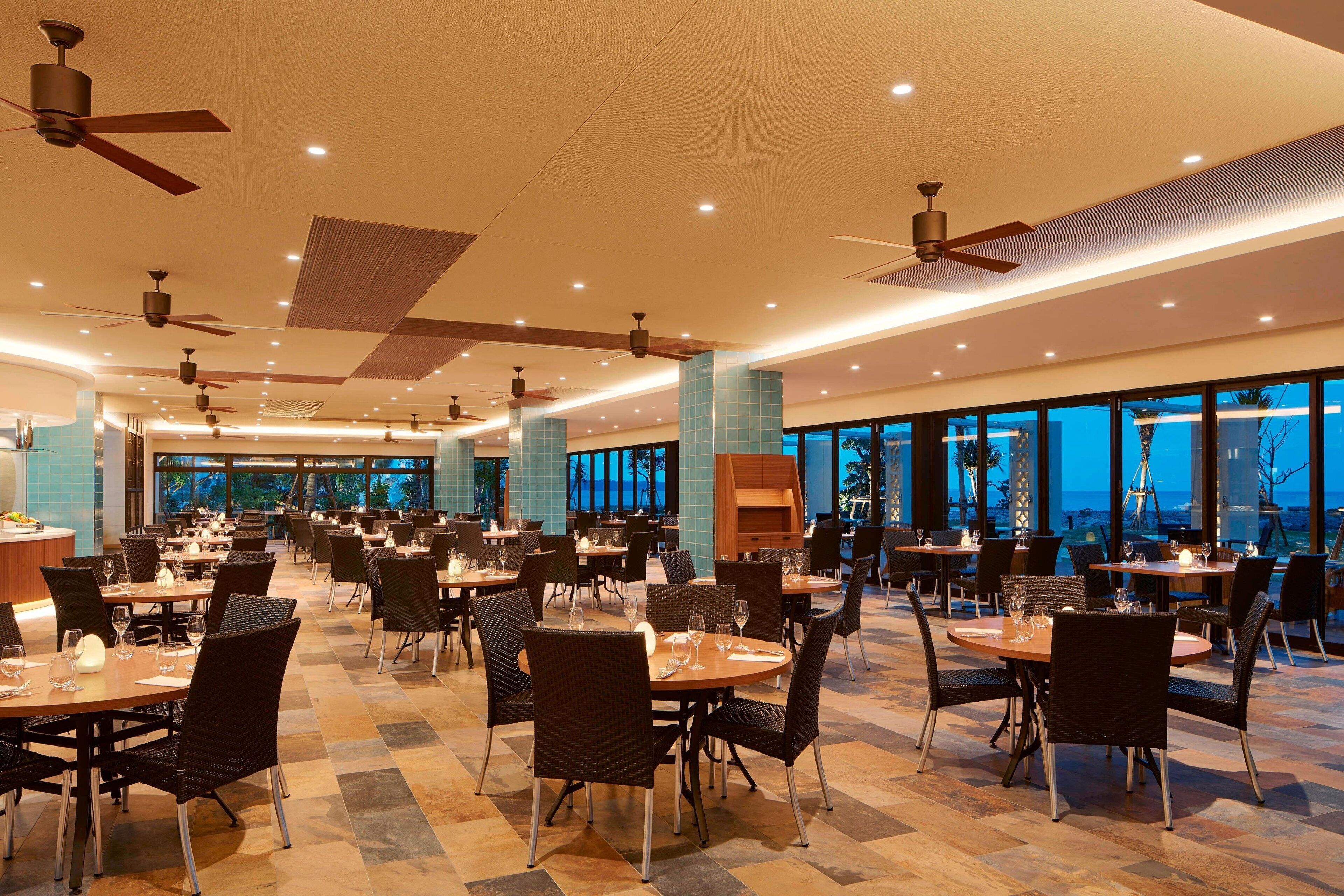 Un vaste espace dédié à la salle à manger, avec de nombreuses tables et chaises, des ventilateurs de plafond et de grandes fenêtres offrant une vue sur l’océan.