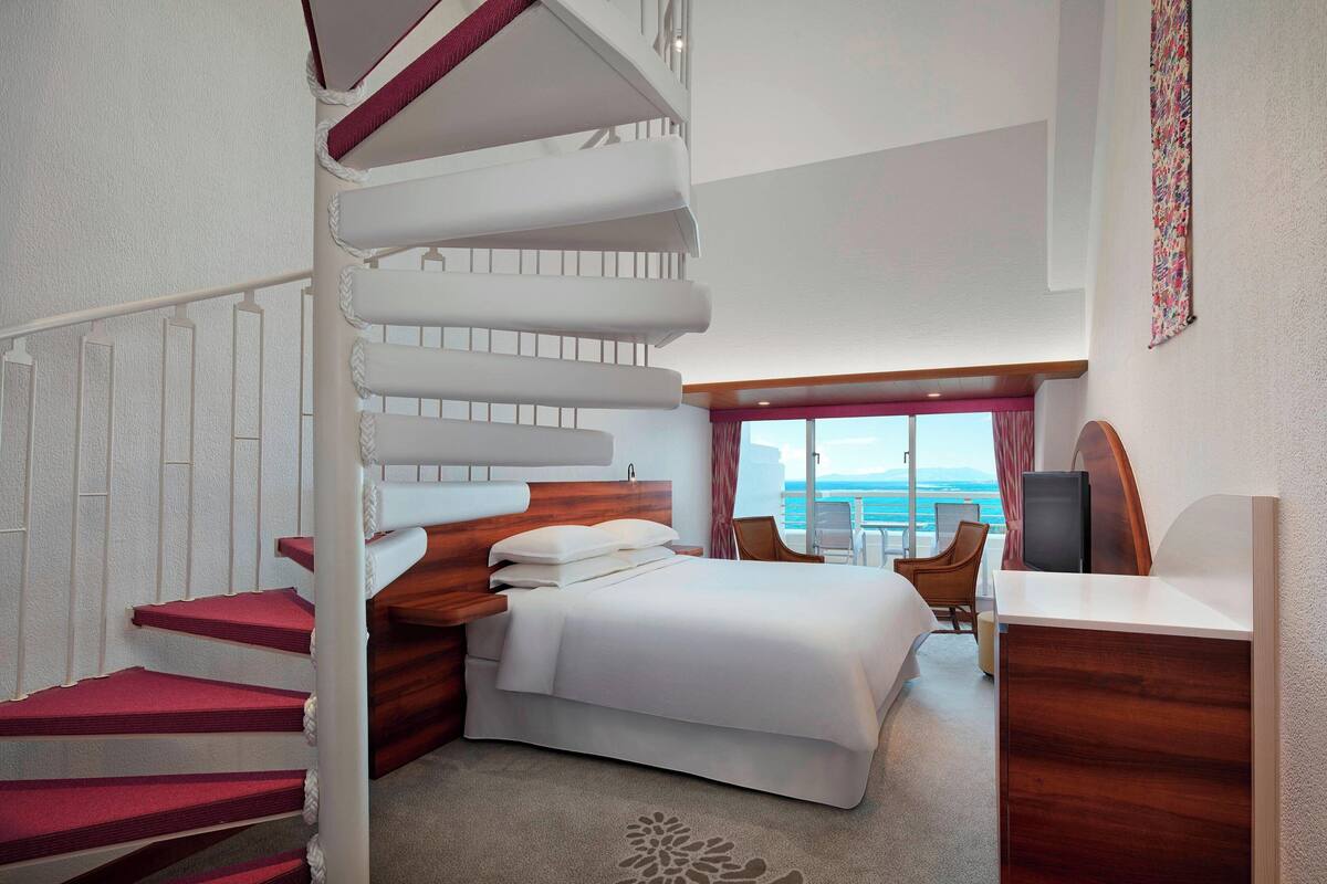 A hotel room with a wooden staircase, a bed with white linens, a desk with a flat-screen TV, and a view of the ocean through large windows.