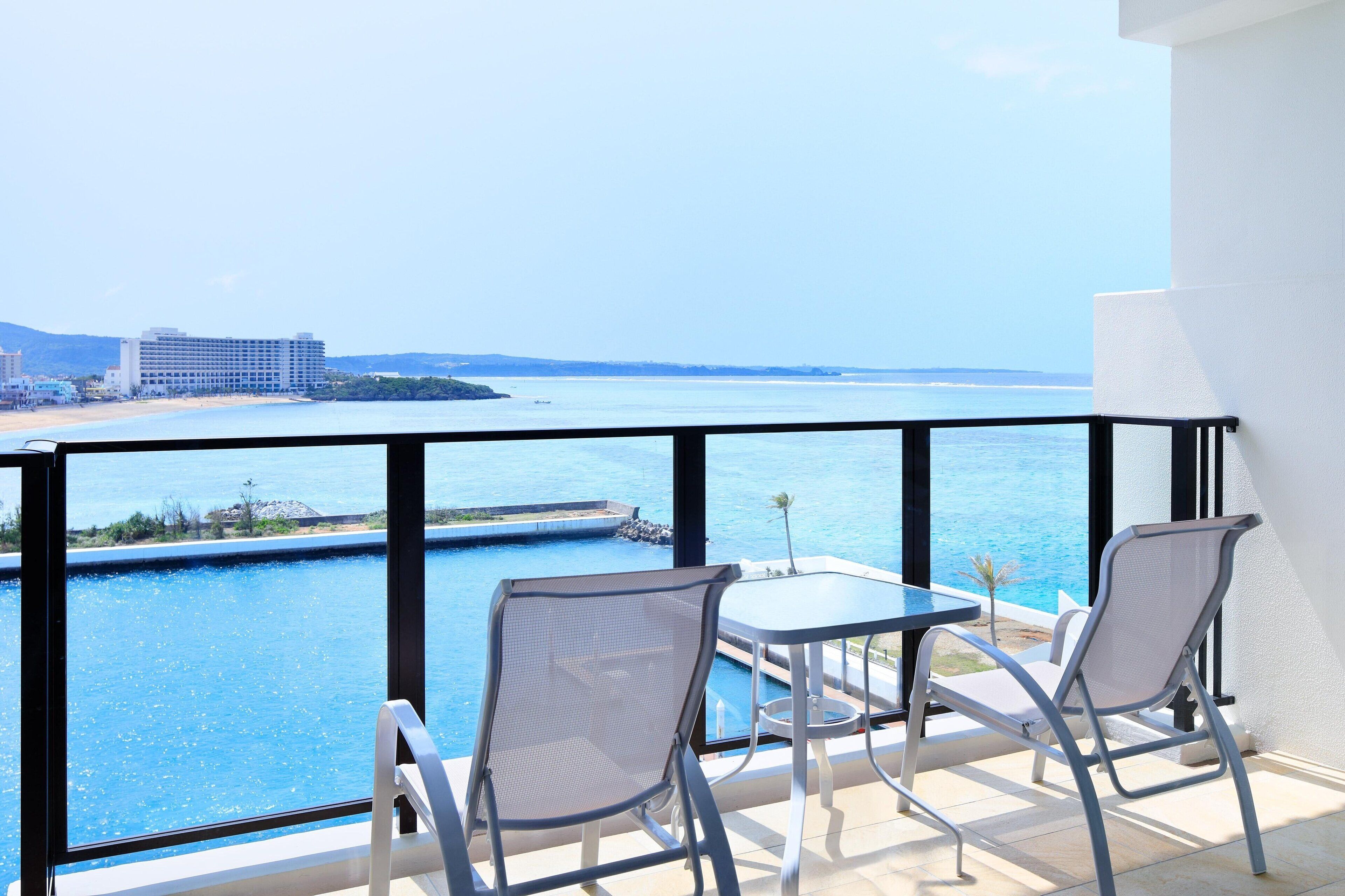 A balcony with two chairs and a table overlooking a swimming pool and a beach.