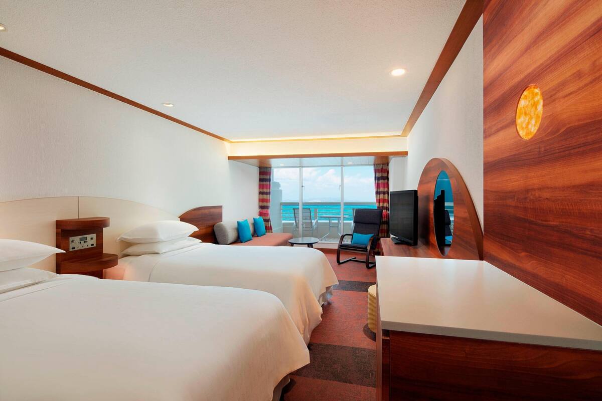 A hotel room with two beds, a TV, a desk, and a view of the ocean.