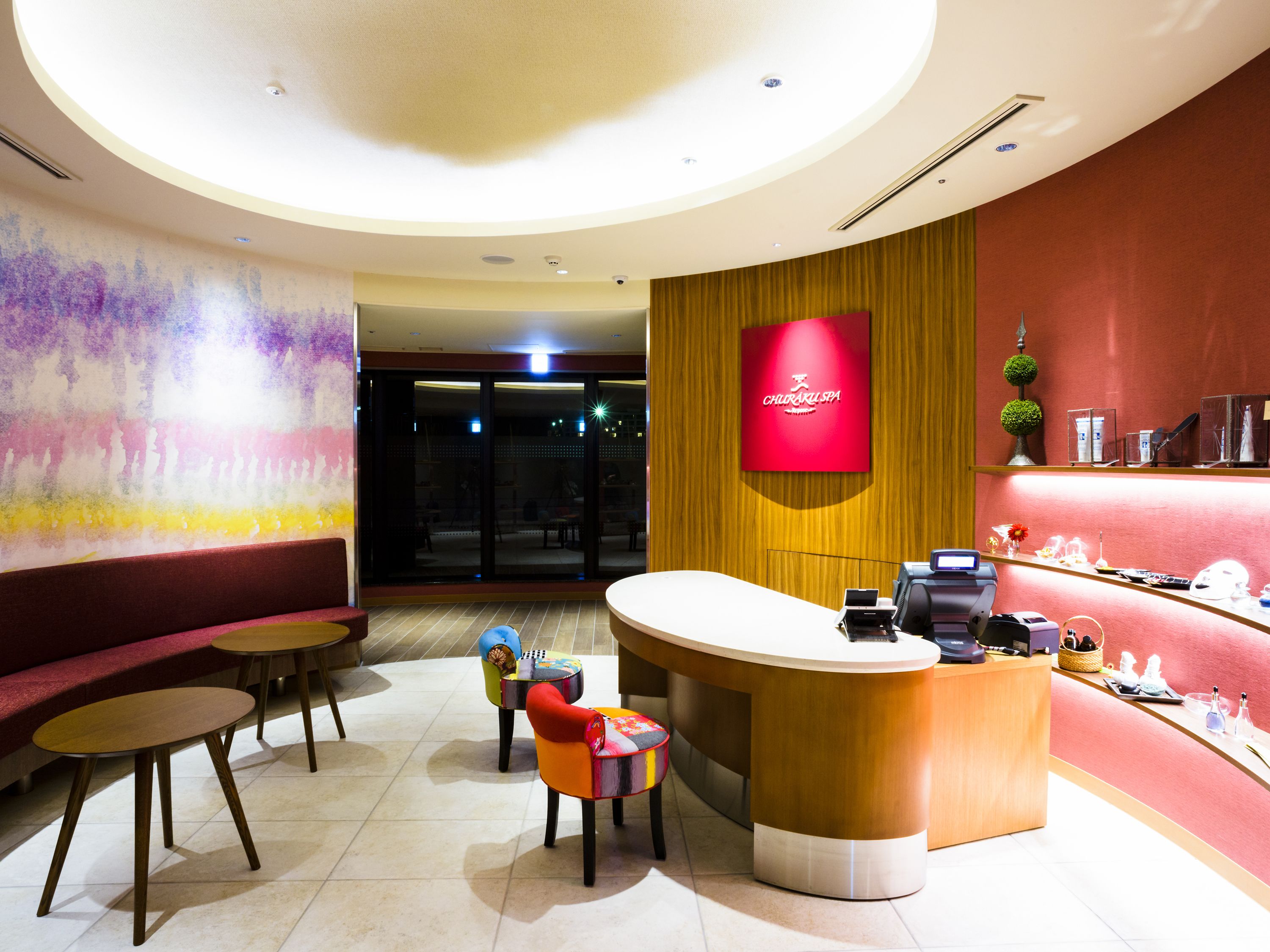 A reception area with a curved counter, a red wall with a spa logo, a seating area with a colorful mural, and a wooden panel wall.