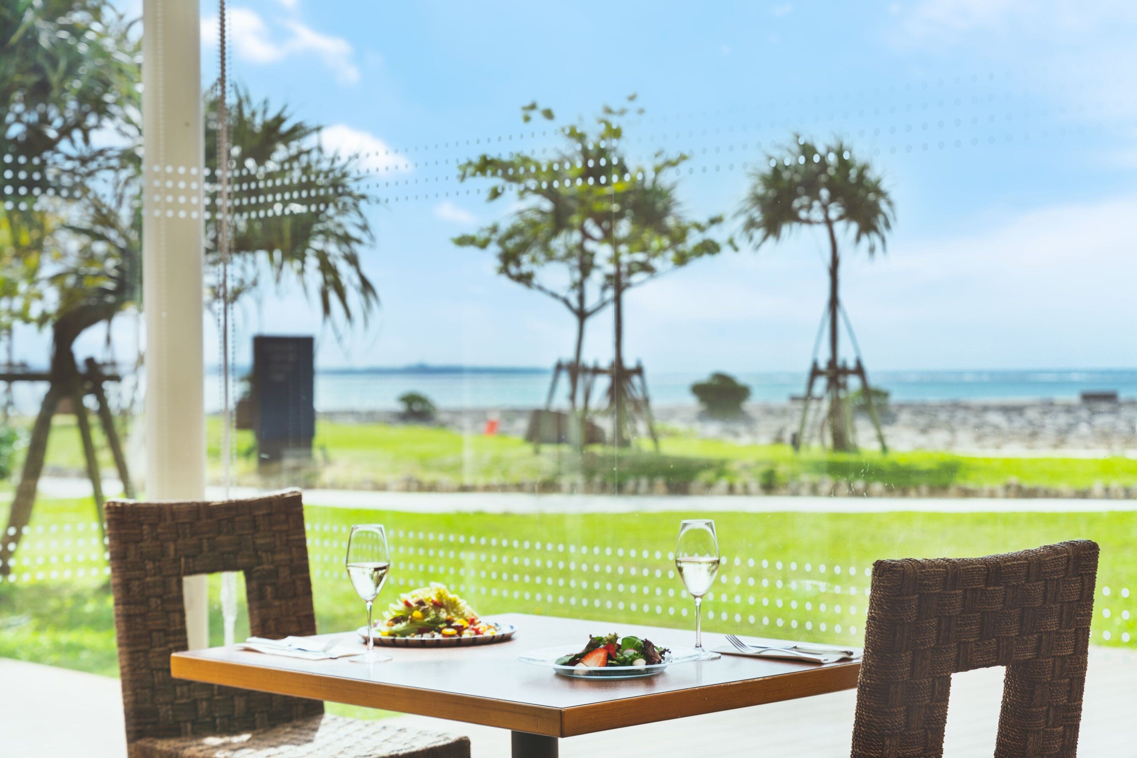 A dining table with two chairs, plates of food, and wine glasses. The table is set outdoors with a view of the beach and palm trees.