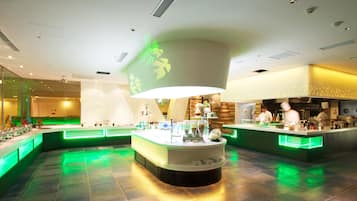 A modern restaurant interior with a central island, green ambient lighting, and a kitchen area visible in the background.