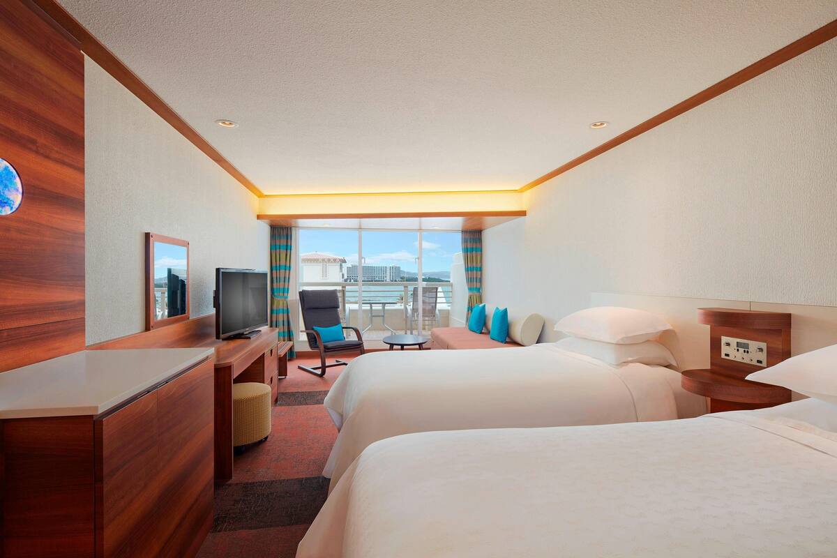 A hotel room with two beds, a desk, a TV, and a balcony with a view of the city.