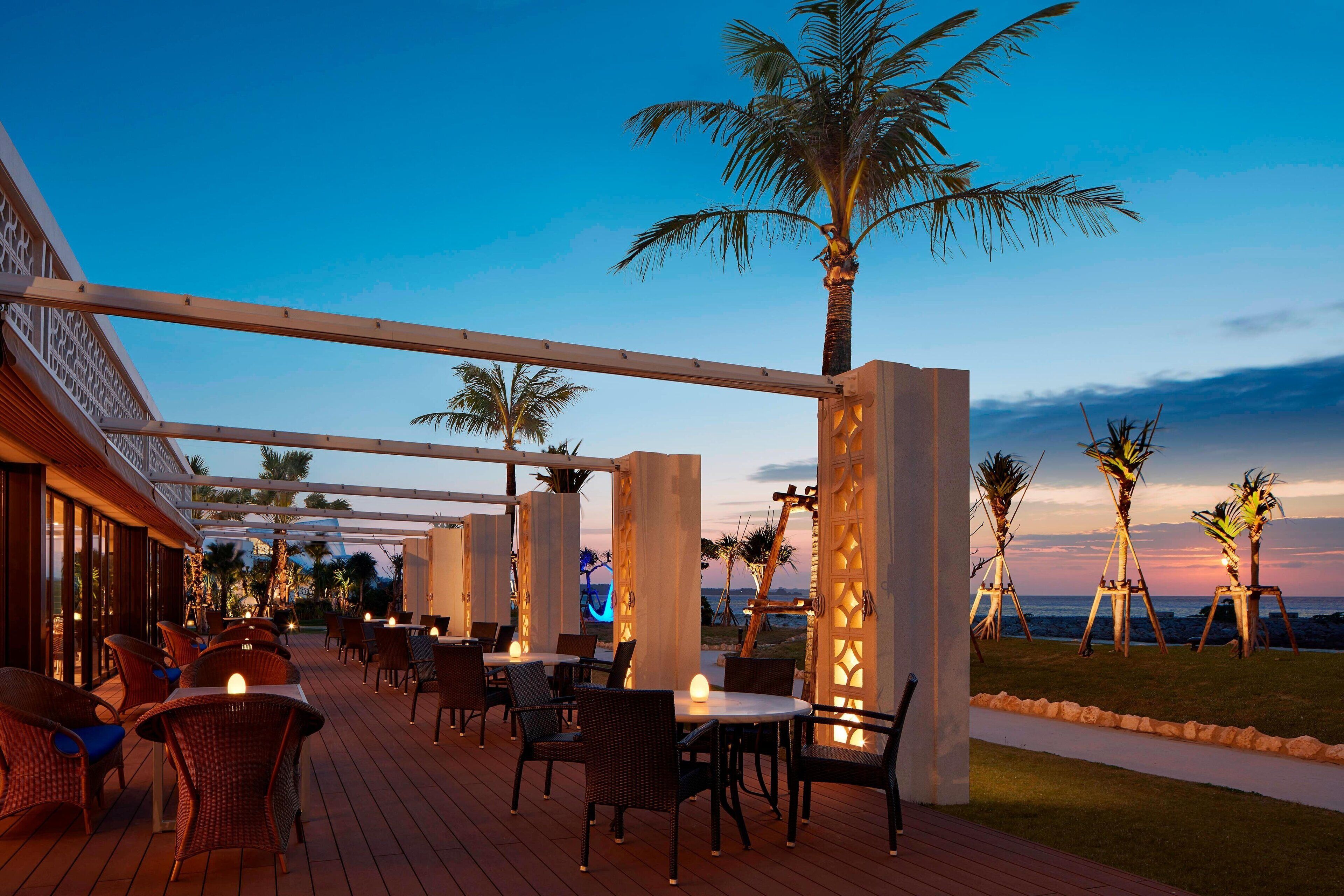 An outdoor patio with tables and chairs, palm trees, and a view of the ocean at dusk.