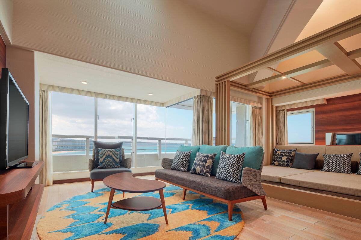 A spacious living area with a sofa, armchairs, a coffee table, and a television. Large windows offer a view of the ocean.