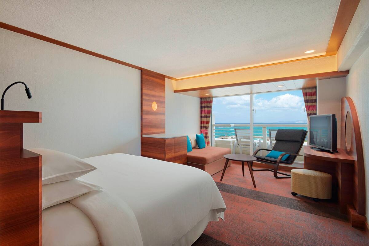 A hotel room with a bed, a TV, a seating area with a view of the ocean, and a window with curtains.