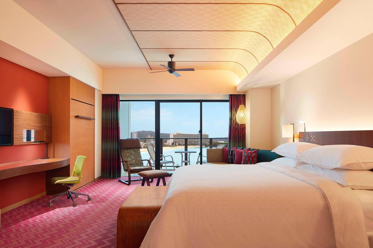 A hotel room with a bed, desk, and seating area. It features a large window with a view of the beach and a ceiling fan.
