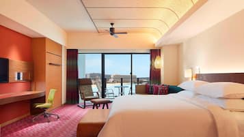 A hotel room with a bed, desk, and seating area. It features a large window with a view of the beach and a ceiling fan.