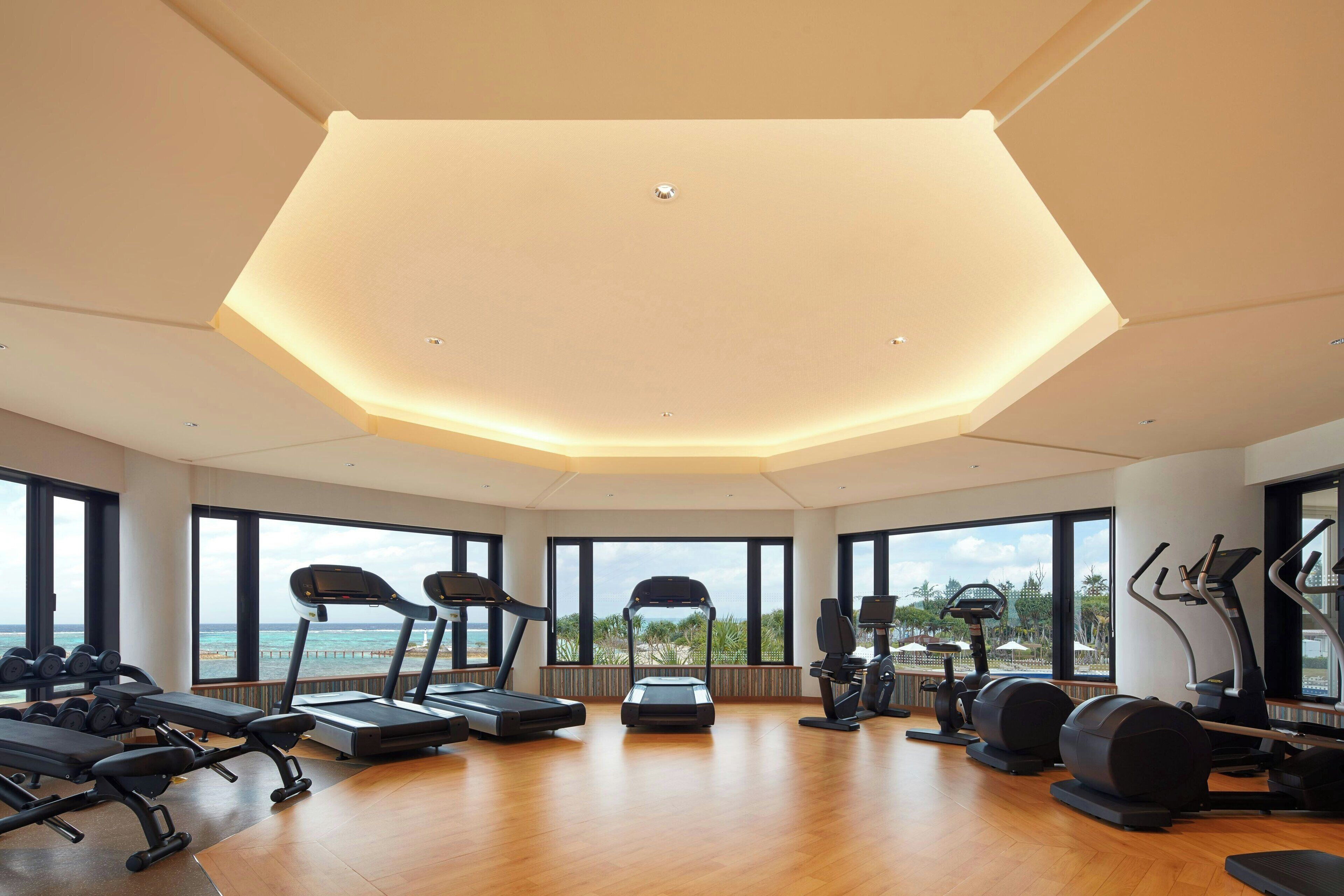 Un gimnasio amplio con diversos equipos de ejercicio, amplias ventanas que ofrecen vistas al océano y un diseño de techo moderno.