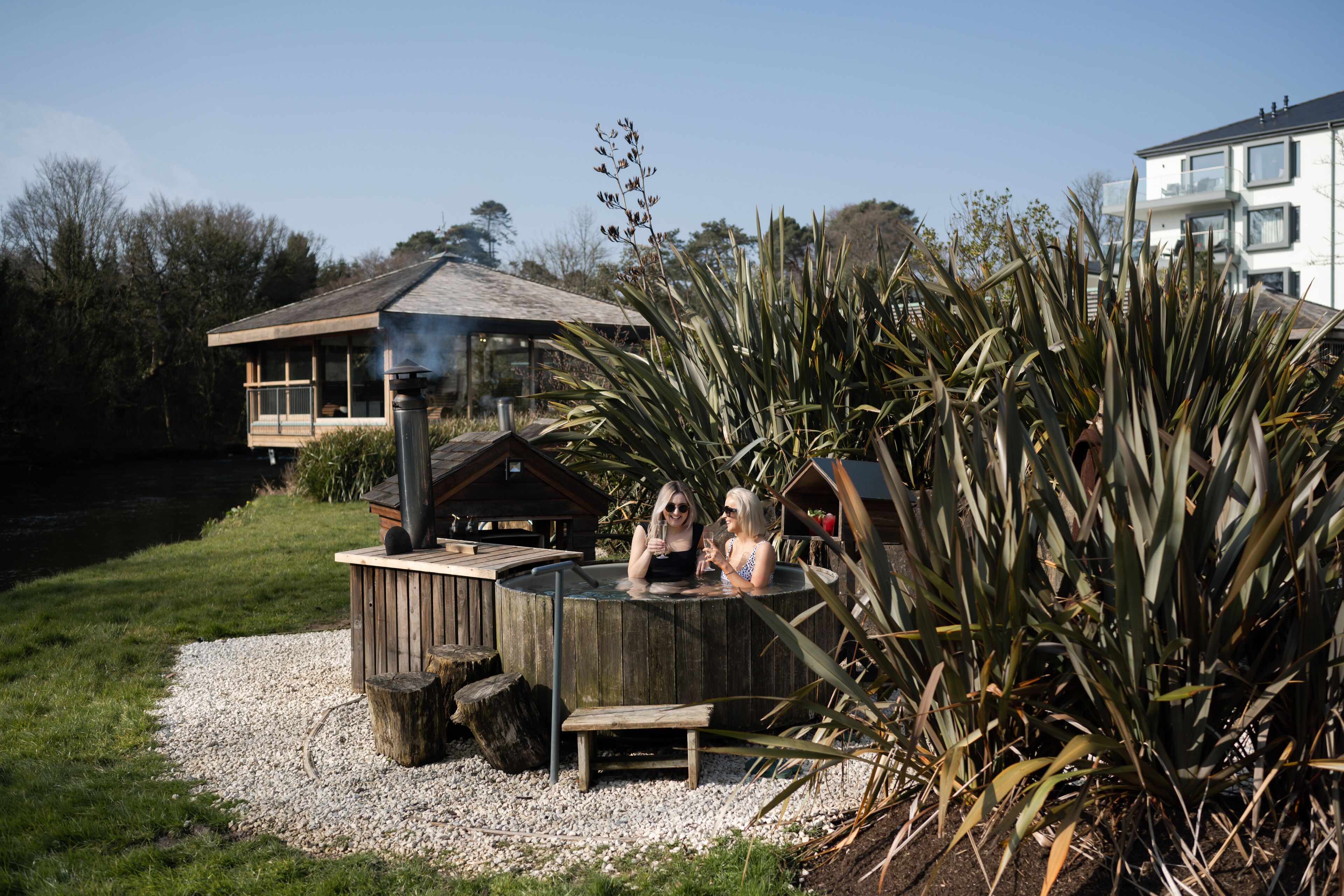 Un jacuzzi con estructura de madera, rodeado de plantas, con un edificio al fondo.