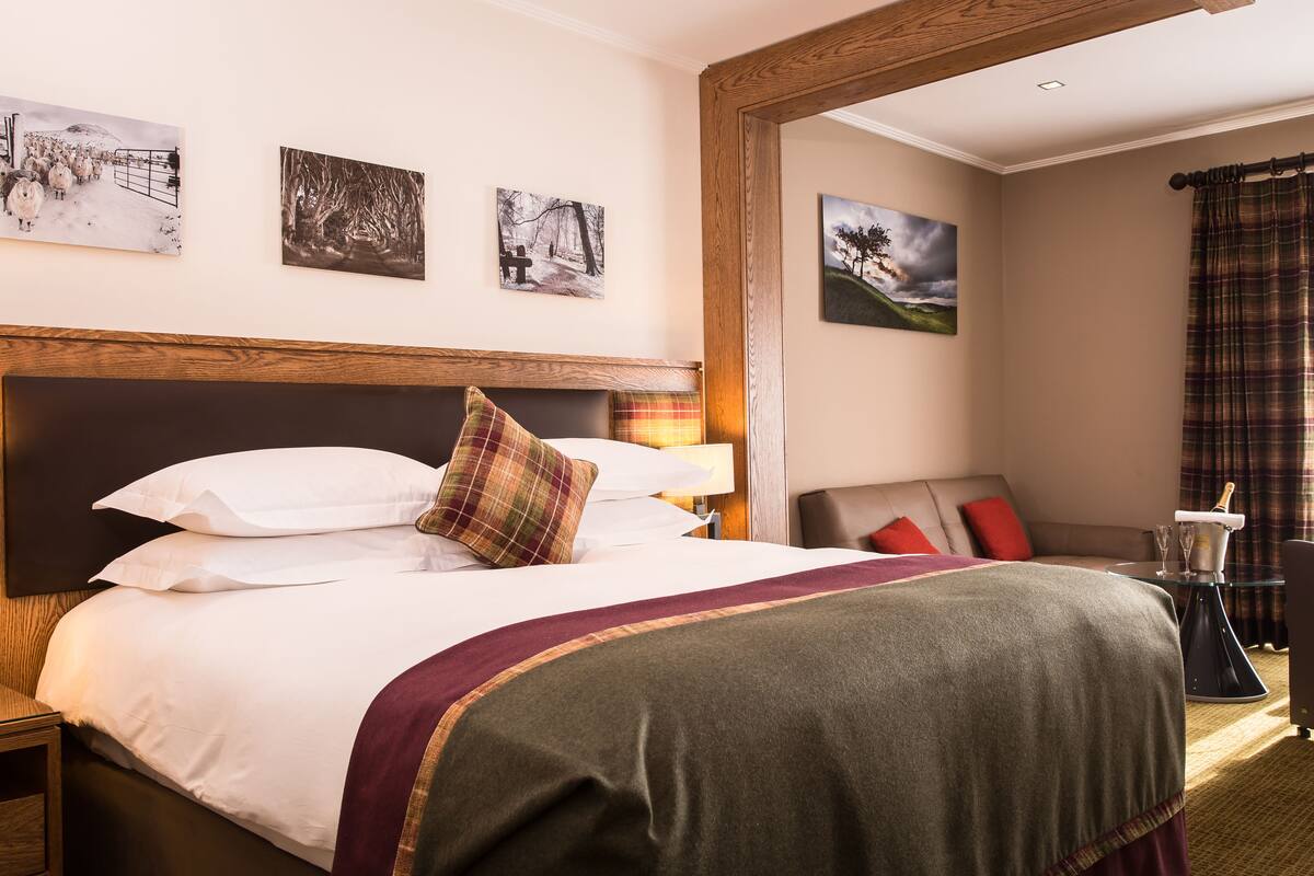 A hotel room with a large bed, a wooden headboard, and a nightstand. There are three paintings on the wall, a window with curtains, and a small table with a lamp and a bottle.