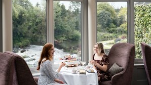 A conservatory with a glass ceiling, large windows, and a table set for two with a view of a river and greenery. - Galgorm (Ballymena)