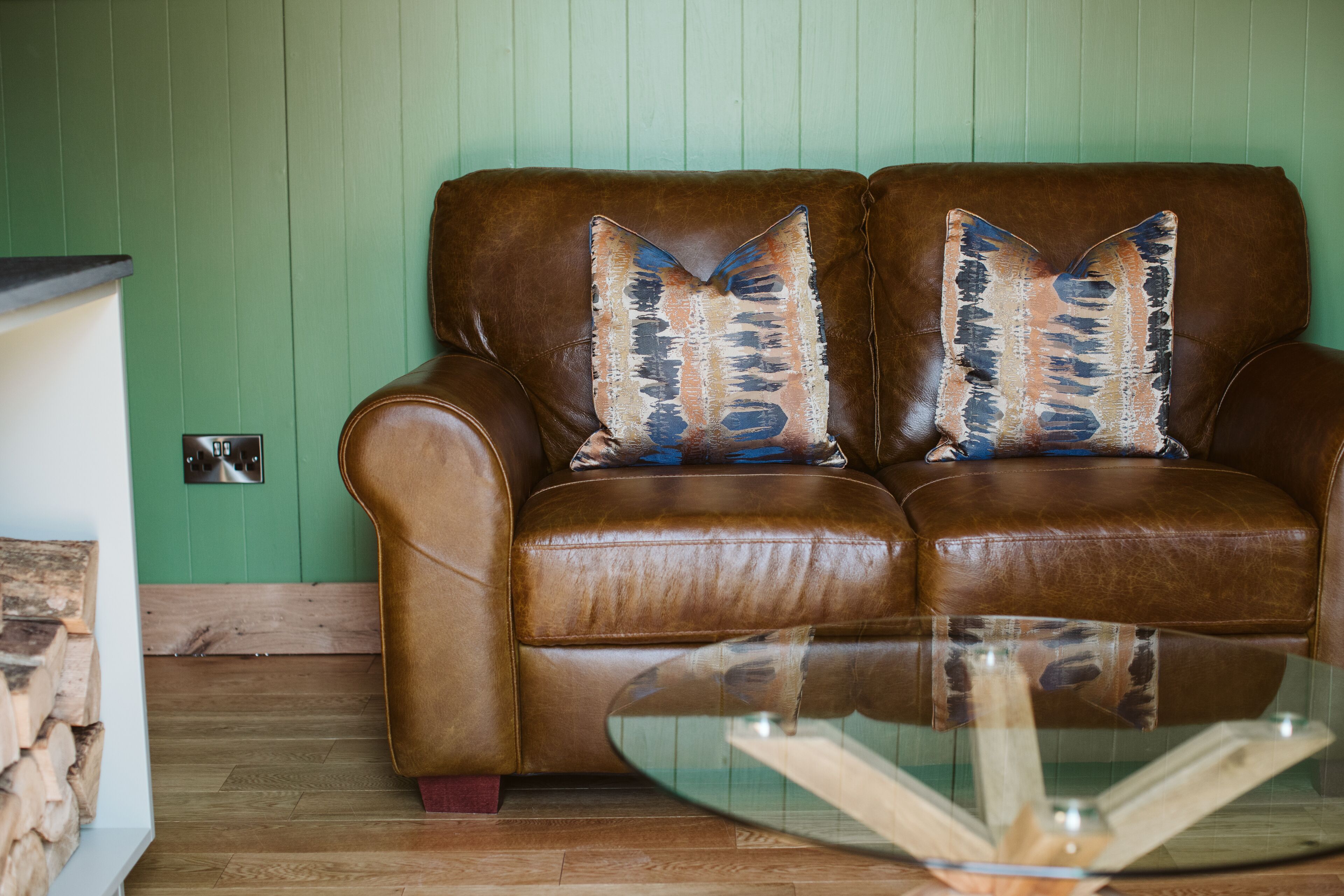 A brown leather sofa with two decorative pillows against a green wall, a glass coffee table in front, and a stack of firewood to the left.
