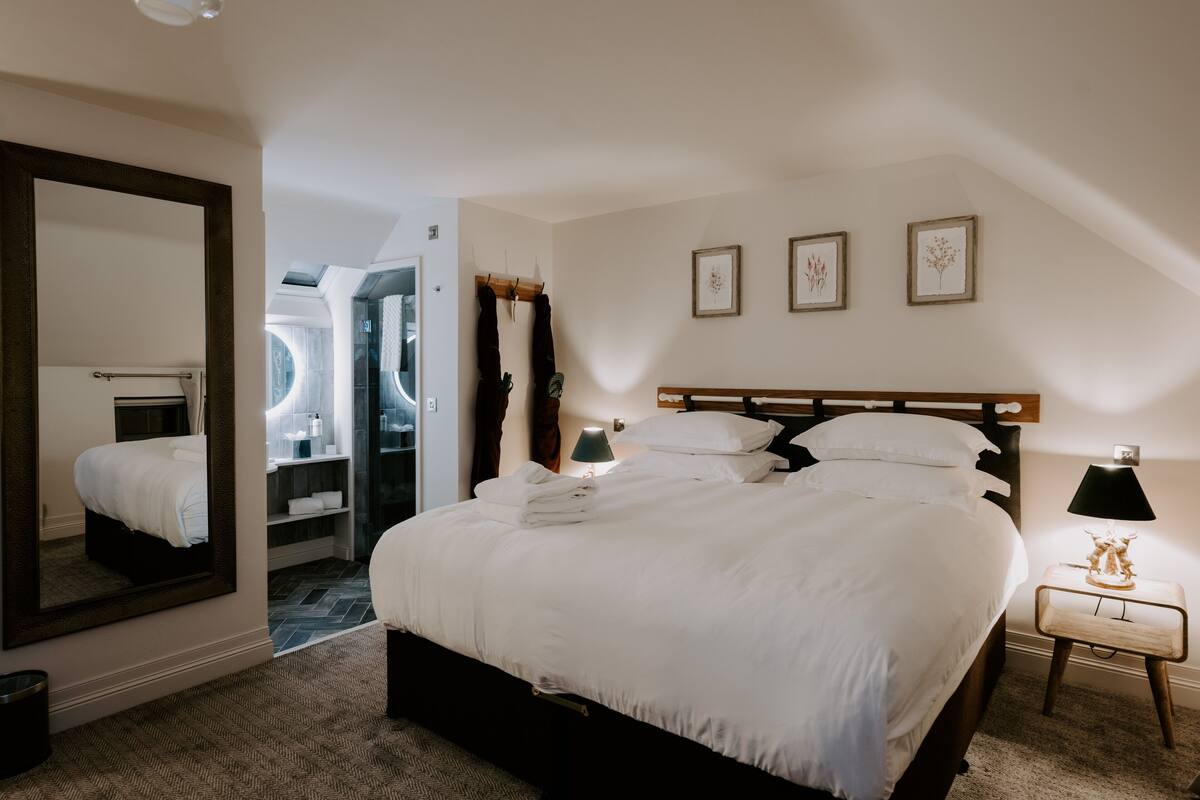 A neatly made bed in a hotel room with a large mirror, a bedside table with a lamp, and framed artwork on the wall.