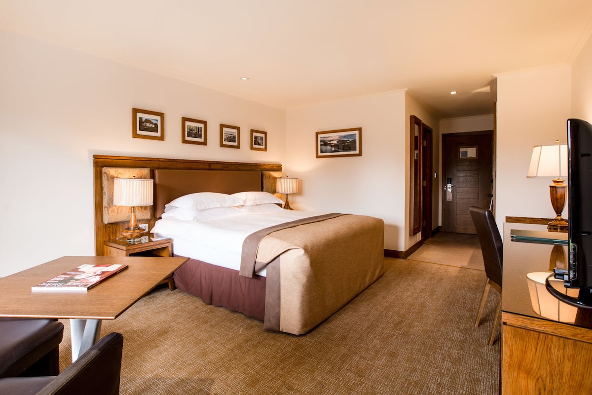 A hotel room with a bed, bedside table, lamp, TV, and a desk with a chair.