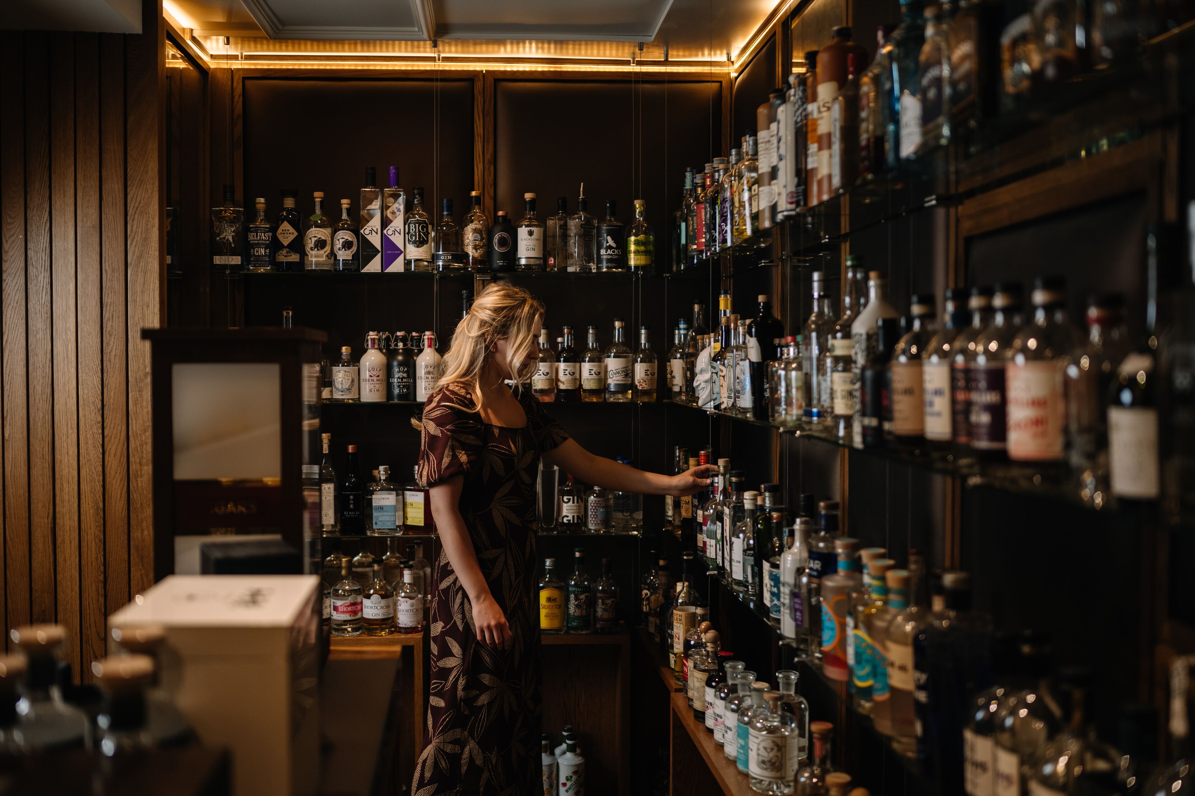 A woman in a dress browsing through a well-stocked liquor shelf in a dimly lit bar.