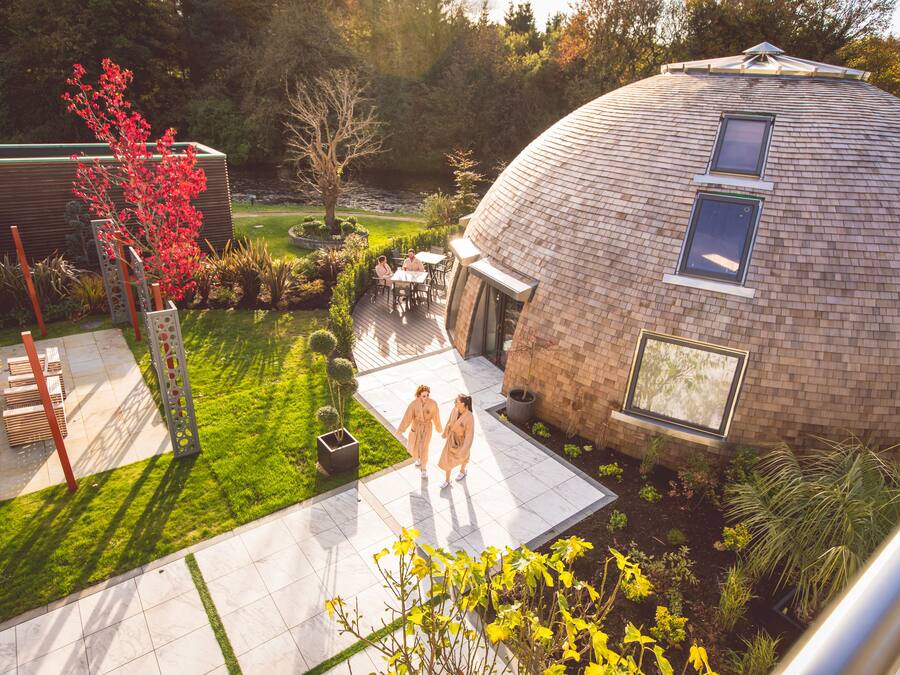 A modern house with a dome-shaped roof, a patio with a table and chairs, and a well-maintained garden with various plants and a red flowering tree.