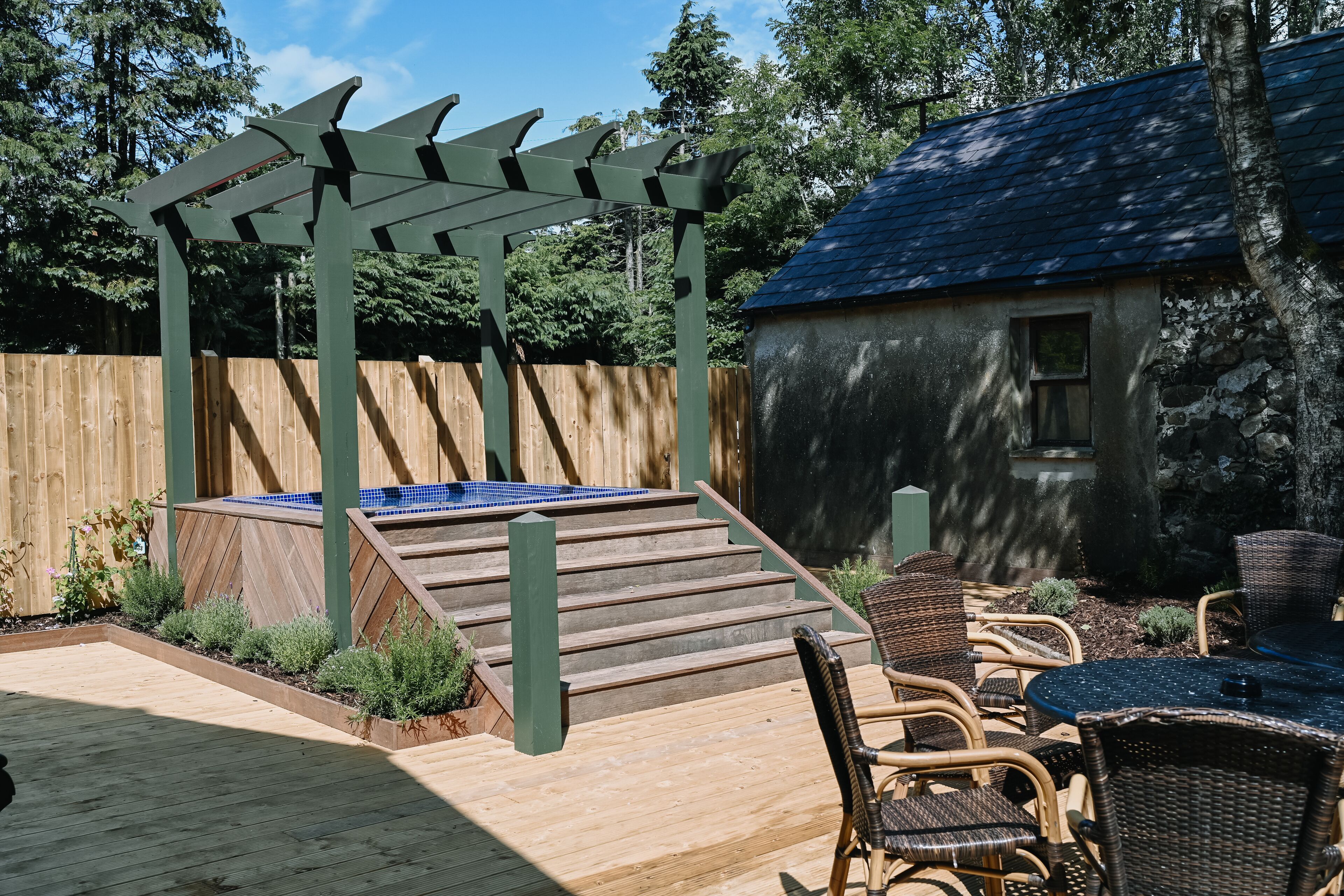 Una terraza de madera con un jacuzzi, sillas de mimbre y una pérgola.