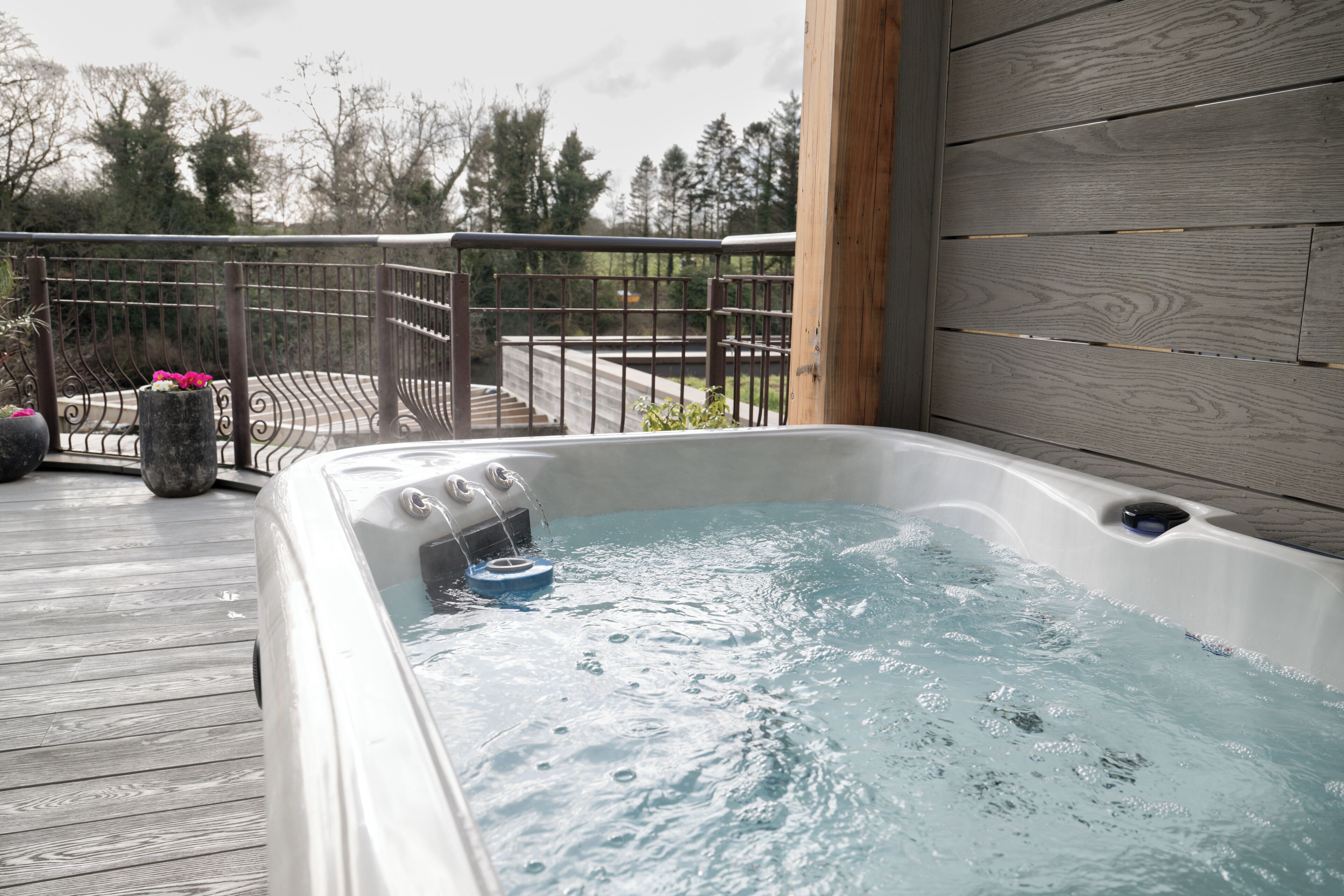 Un bain à remous sur une terrasse en bois, avec une rambarde en métal, entouré d’arbres.