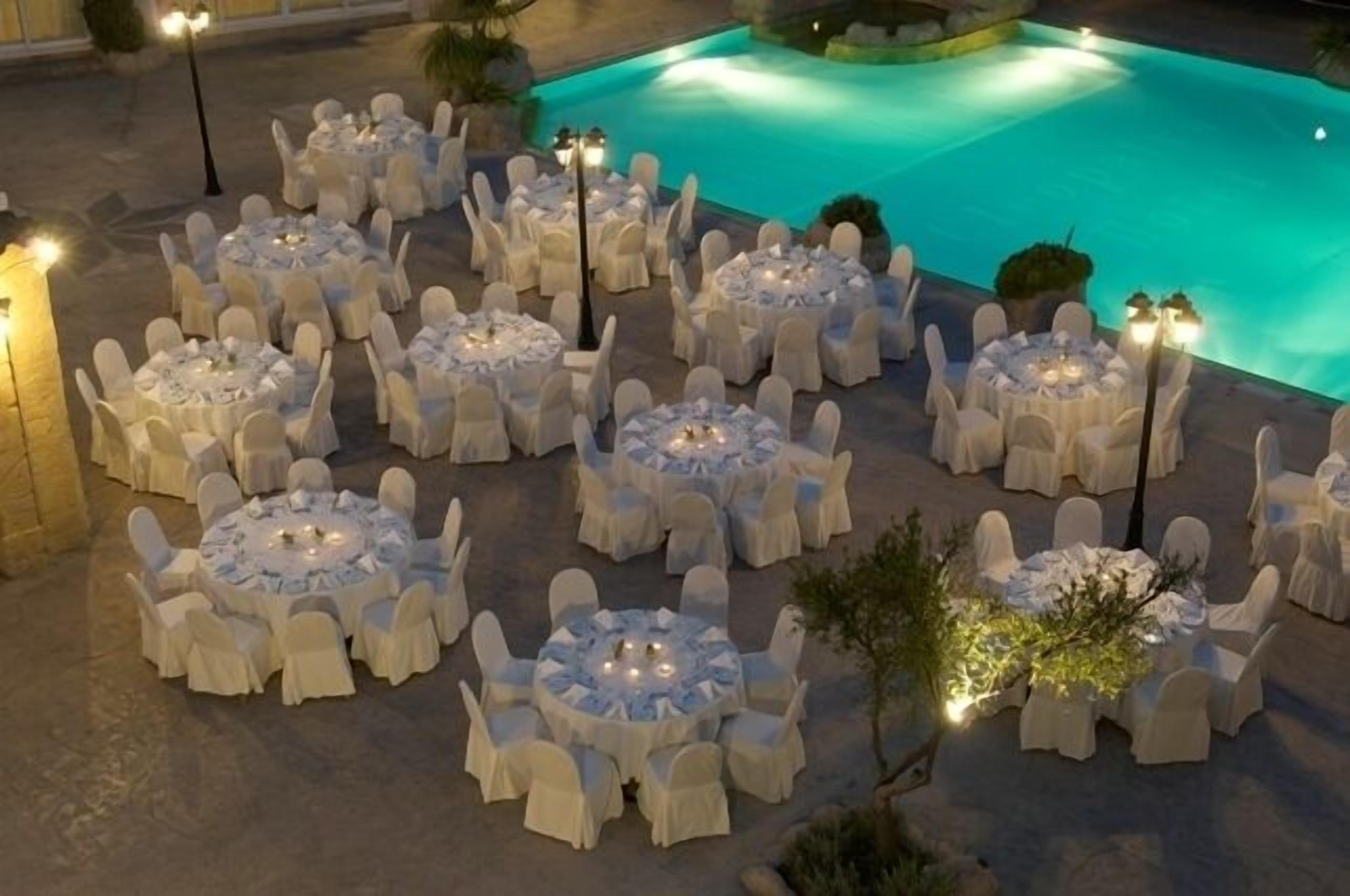 An outdoor dining area with round tables and chairs, illuminated by ambient lighting, near a pool.