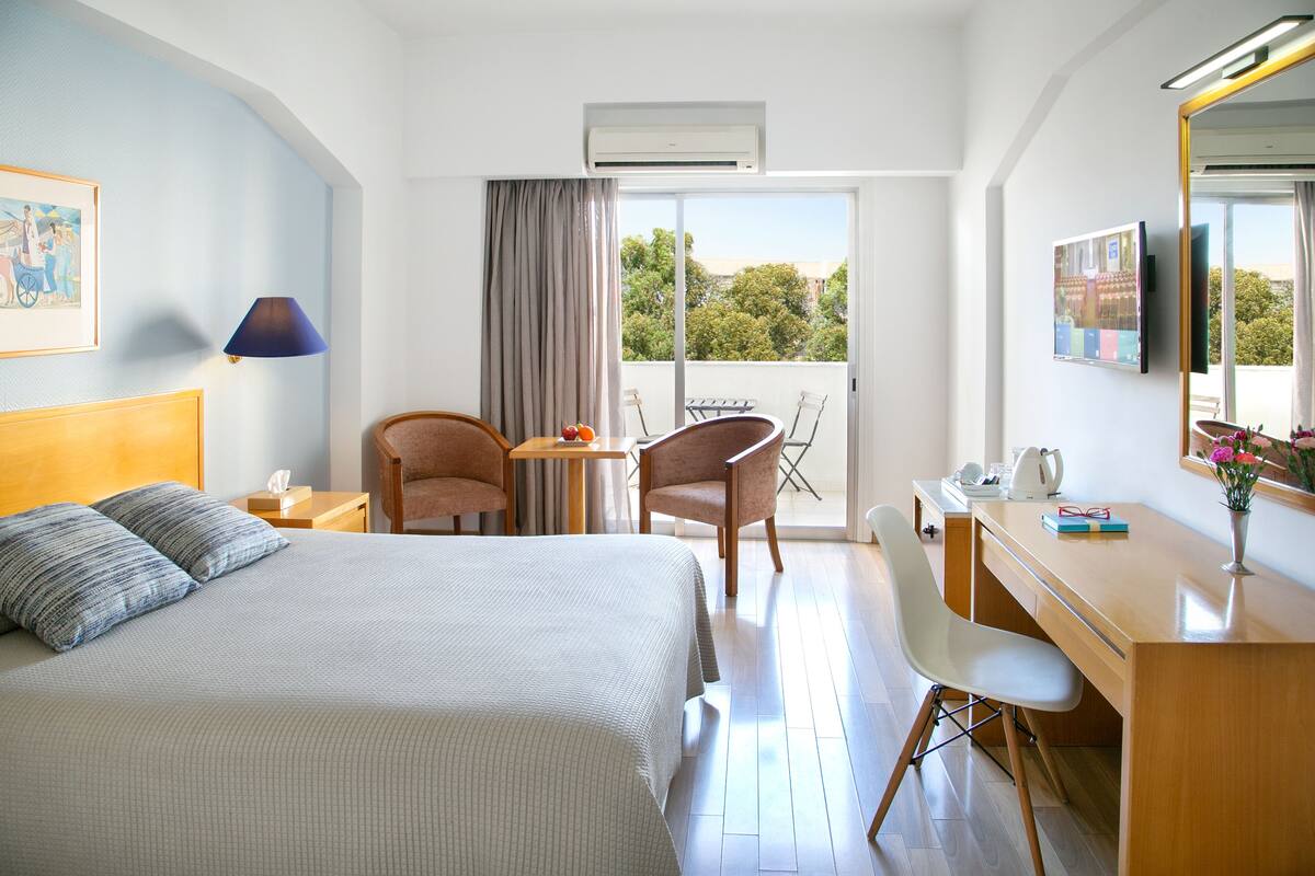 A hotel room with a bed, a desk, a chair, a table, and a balcony with a view of trees.
