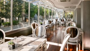 A restaurant with a modern interior, featuring white chairs and tables, large windows, and a view of the street outside. - Cleopatra Hotel (Nicosia)