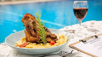 A plate of pasta with a roasted meat and rosemary garnish, a glass of red wine, and a menu on a table by a pool.