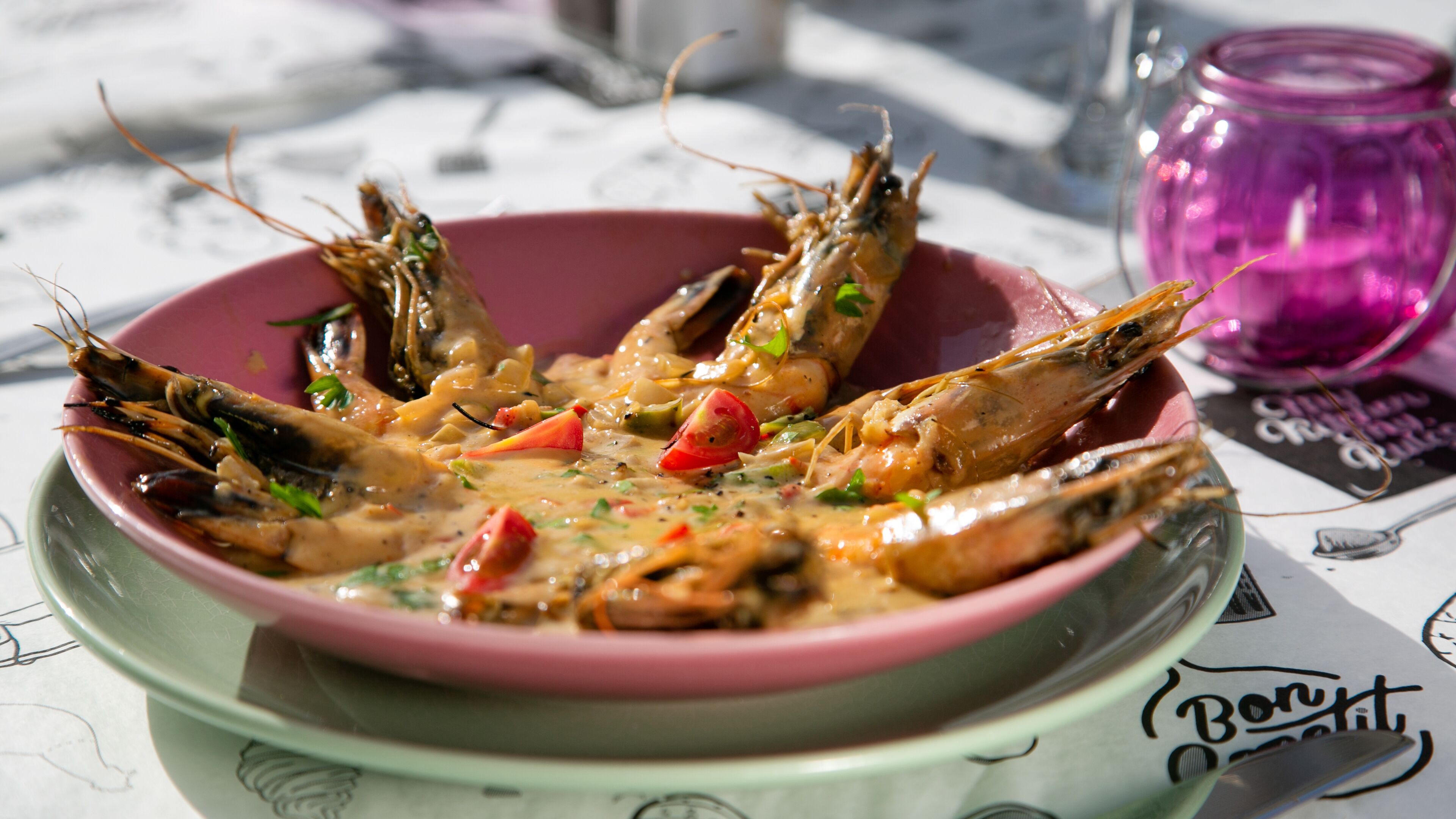 A bowl of cooked shrimp with vegetables and sauce on a plate, with a pink glass and a menu in the background.