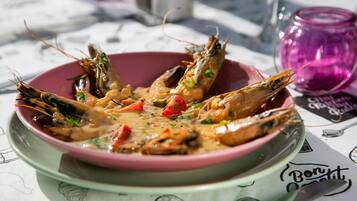 A bowl of cooked shrimp with vegetables and sauce on a plate, with a pink glass and a menu in the background.