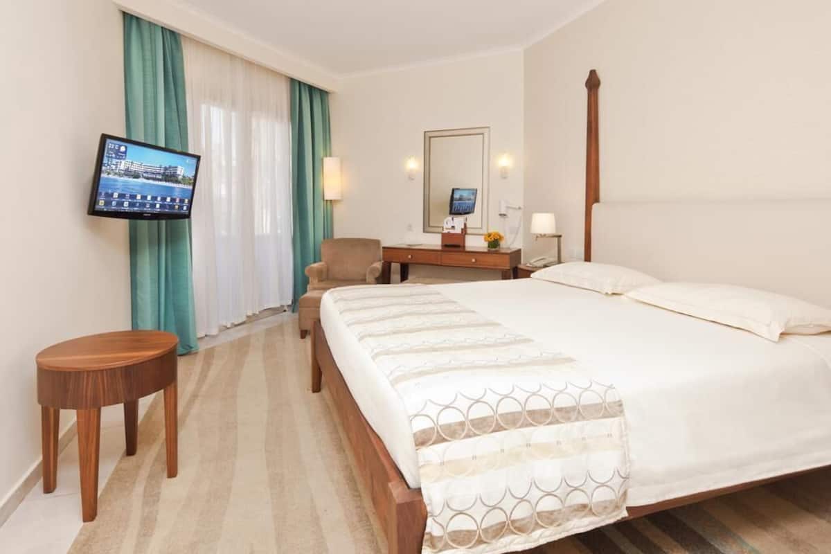 Ein Hotelzimmer mit Bett, Schreibtisch, Sessel, Fernseher und einem fenster mit Vorhängen.