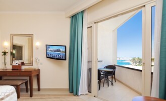 Habitación de hotel con balcón, escritorio, silla, televisor y vista al mar.