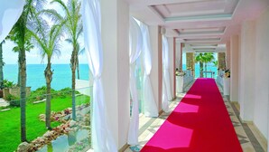 Un couloir d’hôtel avec un tapis rouge menant à un balcon d’où l’on peut admirer la mer, les palmiers et un ciel dégagé.