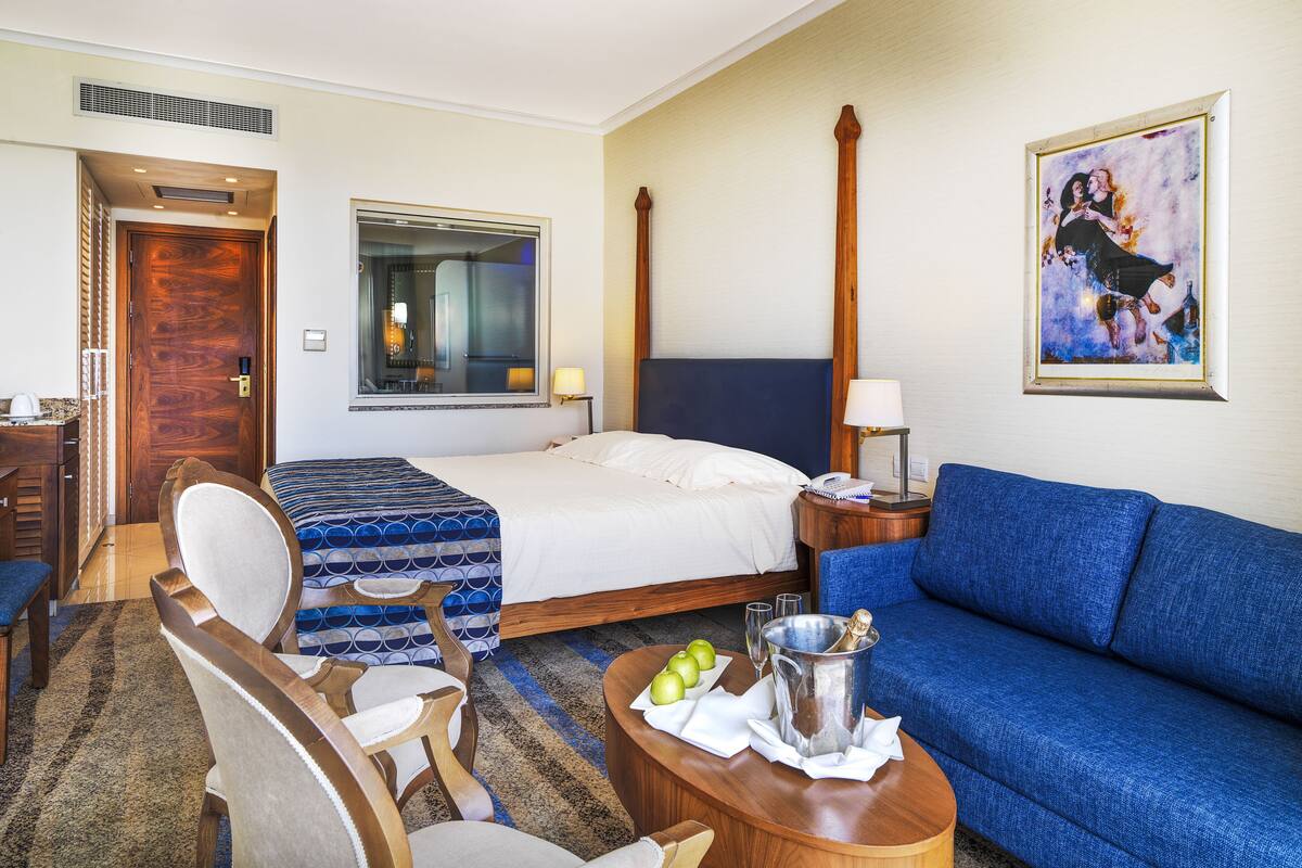 A hotel room with a bed, a blue sofa, a wooden side table with a bottle and glasses, a chair, and a painting on the wall.