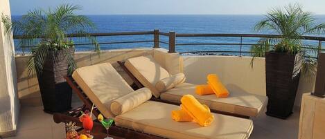 A sun lounger with cushions and towels, a small table with fruit and drinks, and potted plants on a balcony with a view of the sea.
